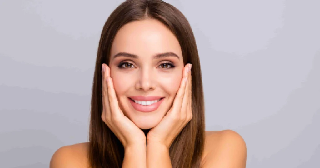 A smiling woman with long brown hair and natural makeup gently touches her cheeks, showing smooth, radiant skin during Daxxify® Treatments in Kirkland, WA.