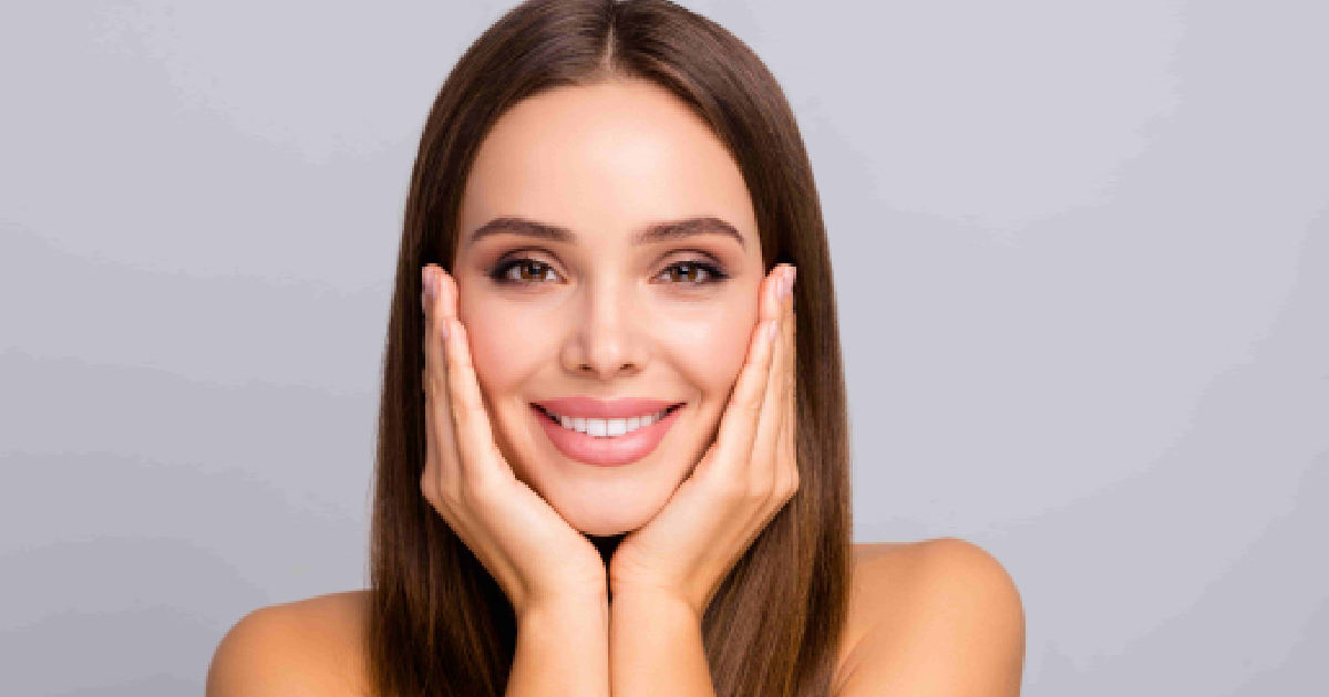 A smiling woman with long brown hair and natural makeup gently touches her cheeks, showing smooth, radiant skin during Daxxify® Treatments in Kirkland, WA.