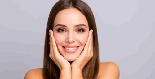 A smiling woman with long brown hair and natural makeup gently touches her cheeks, showing smooth, radiant skin during Daxxify® Treatments in Kirkland, WA.